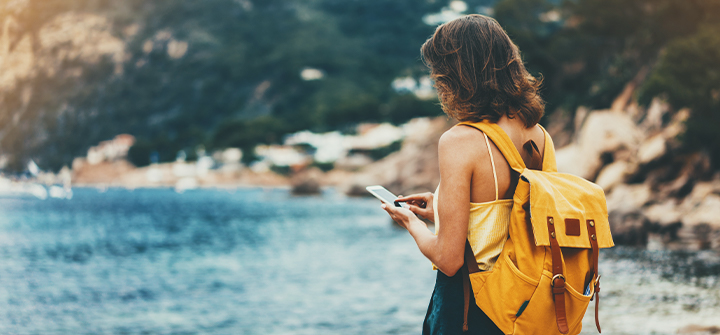 Mulher em frente ao mar utilizando o celular e com mochila amarela